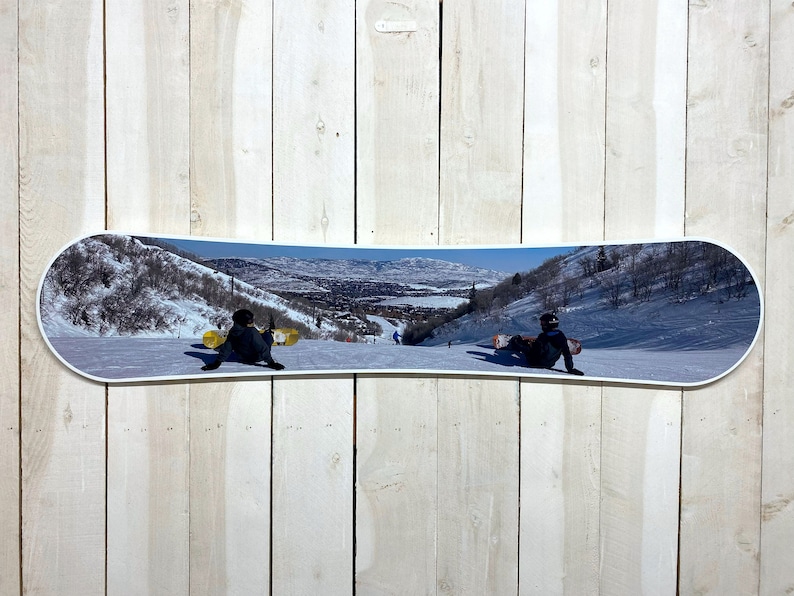Puede incluir: Un snowboard con una foto panor&aacute;mica de un paisaje de monta&ntilde;a nevado. Dos personas est&aacute;n haciendo snowboard bajando la monta&ntilde;a, de espaldas a la c&aacute;mara.