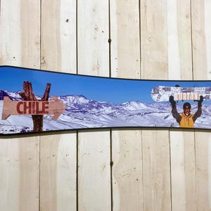 Puede incluir: Un snowboard con una escena de monta&ntilde;a azul, blanca y marr&oacute;n. El snowboard tiene un marco negro y un cartel que dice "CHILE" con una flecha apuntando hacia la derecha. Una persona est&aacute; sosteniendo una tabla de snowboard por encima de su cabeza.