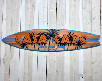 Pode incluir: Uma placa de madeira de prancha de surf com o texto "Casa Cayo" em letras laranja e o texto "Life is Good Today" em letras azuis. A placa é decorada com palmeiras e tem um design de listras azuis e brancas.