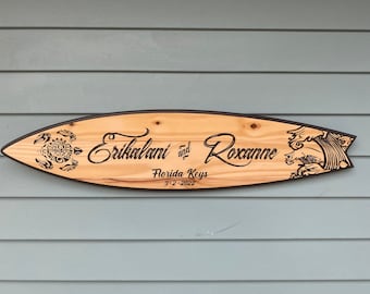 May include: A surfboard-shaped wooden wall sign with a natural light wood grain finish and dark brown edges. The sign features a black tribal-style sea turtle, the text "Erikalani and Roxanne", "Florida Keys", and the date "3-2-2022", plus a wave graphic.