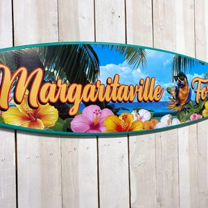 May include: A surfboard-shaped sign with the words "Margaritaville Forever" in orange and yellow lettering. The sign features a tropical scene with a parrot, flowers, palm trees, and a blue ocean background.