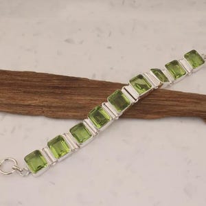 Pode incluir: Uma pulseira de prata com pedras preciosas retangulares verde claro. As pedras são engastadas em molduras de prata individuais, conectadas por elos de prata. A pulseira é exibida em um pedaço de madeira sobre um fundo branco. Possui um fecho de alternância.