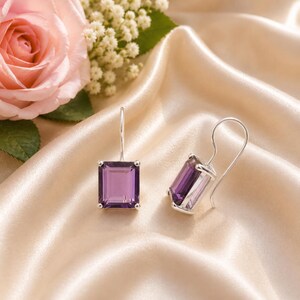 May include: A pair of amethyst earrings with a rectangular cut. The purple gemstones are set in silver-colored metal. The earrings are displayed on a satin surface, with a pink rose and white flowers in the background.