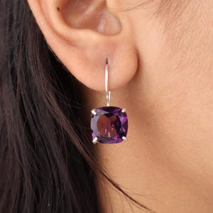May include: A close-up of a single earring with a square, deep purple gemstone. The gemstone is set in a silver metal frame and hangs from a silver hook. The earring is worn on a person's ear, with their dark hair visible.