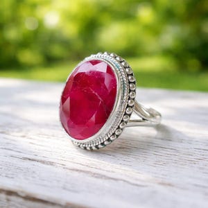 Op de afbeelding: Zilveren ring met een grote, ovaal geslepen robijn. De robijn is dieprood en gezet in een zilveren zetting met een kralenrand. De ring ligt op een wit houten oppervlak met een vage groene achtergrond.