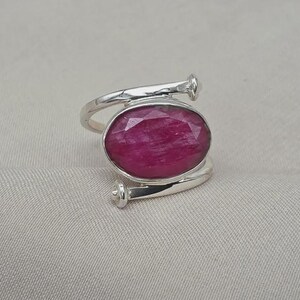 Op de afbeelding: Een zilveren ring met een grote, ovaalvormige, gefacetteerde dieproze robijn in het midden. De ringband is ontworpen als een metalen spijker die om de vinger wikkelt, waarbij de kop van de spijker aan beide uiteinden van de ring zichtbaar is.