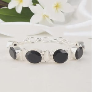 Peut inclure: Un bracelet en argent orné de pierres noires ovales. Le bracelet présente un design à maillons et est posé sur un fond blanc avec des fleurs blanches.