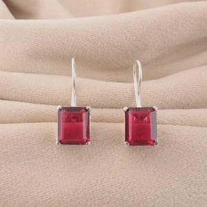 May include: A pair of silver drop earrings with rectangular, deep red gemstones. The earrings have a simple, elegant design, with the gemstones set in a silver frame and suspended from a curved hook.