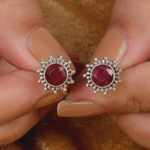 Può includere: Un paio di orecchini a lobo con pietre preziose rotonde e sfaccettate di colore rosso intenso incastonate in argento. Ogni orecchino presenta una cornice a raggiera con piccole perline d'argento che si irradiano dalla pietra centrale.