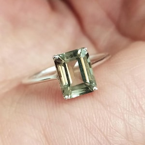 Pode incluir: Um anel de prata com uma pedra preciosa retangular verde claro. A pedra é engastada num design simples e elegante, realçando a sua clareza e cor. A banda do anel é fina e polida.