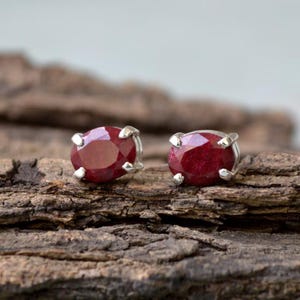 Puede incluir: Un par de pendientes de botón de rubí. Cada pendiente presenta una gema de color rojo intenso, de forma ovalada, engastada en un metal plateado. Los pendientes se exhiben sobre un trozo de madera marrón texturizada.