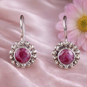 Pode incluir: Um par de brincos pendentes de prata com uma pedra preciosa circular facetada de cor vermelho-escura ao centro. A pedra é rodeada por uma borda decorativa de pequenas esferas de prata e cada brinco tem um gancho de prata curvo.