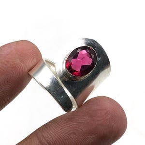 May include: A silver ring featuring a large, oval-cut, magenta gemstone. The ring has a wide, smooth band that curves around the finger. The gemstone is set in a bezel setting, and the ring is shown against a white background.