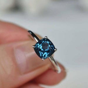 May include: A silver ring with a square, teal-coloured gemstone. The gemstone is held by four small prongs. The ring's band is thin and polished, creating a classic and elegant appearance.