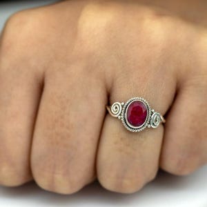 Könnte beinhalten: Ein Silberring mit einem großen, ovalen Rubin-Edelstein. Der Rubin ist in einer strukturierten, seilartigen Lünette gefasst. Der Ring hat dekorative Spiralakzente auf beiden Seiten des Edelsteins.