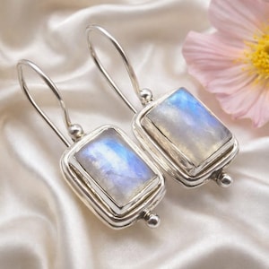 Puede incluir: Pendientes de plata con piedras lunares rectangulares. Las piedras muestran un brillo iridiscente azul y blanco, engastadas en un marco de plata con un diseño de gancho simple. Se presentan sobre una superficie de satén blanco.