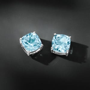 May include: A pair of square-cut, light blue gemstone stud earrings. Each earring features a cushion-cut, pale blue gemstone set in a silver-coloured metal setting. The earrings are displayed on a dark, reflective surface.