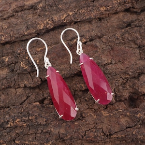 May include: A pair of ruby-coloured gemstone earrings with a faceted, teardrop design. The earrings are suspended from silver-coloured hooks. The gemstones are a deep red hue, and the earrings are displayed on a textured, brown wooden surface.