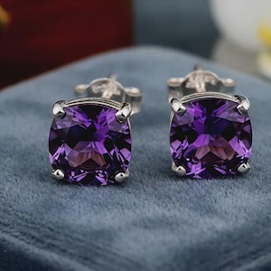 May include: A pair of square-cut amethyst stud earrings. Each earring has a vibrant purple gemstone set in silver-coloured metal. The earrings are displayed on a blue velvet surface, highlighting their colour and brilliance.