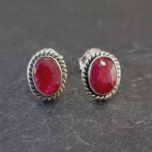 Peut inclure: Une paire de boucles d'oreilles à clous en forme d'ovale avec des rubis. Chaque boucle d'oreille présente une pierre précieuse rouge vif sertie dans un cadre argenté texturé en corde. Les boucles d'oreilles sont présentées sur une surface gris foncé.