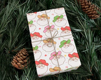 May include: Wrapped gift with a festive pattern of Santa Claus faces wearing cowboy hats in red, green, pink, and tan. The gift is tied with twine and set against a backdrop of pine needles and pine cones.
