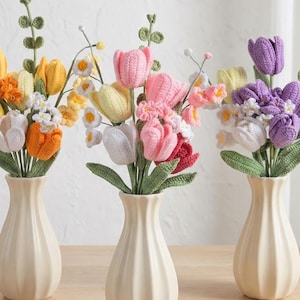 Könnte beinhalten: Drei Vasen mit bunten, gehäkelten Blumen, darunter Tulpen, in Gelb-, Orange-, Rosa-, Rot-, Weiß- und Lilatönen. Die cremefarbenen Vasen stehen auf einer Holzoberfläche. Eine Geschenkbox und eine Kerze sind ebenfalls zu sehen.