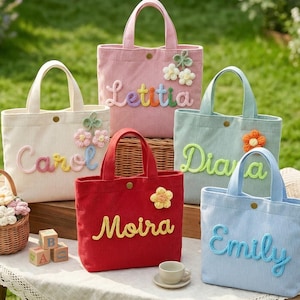 Puede incluir: Una colección de bolsos tote de pana en varios colores pastel, cada uno con un nombre bordado en la parte delantera. Las bolsas están decoradas con adornos florales y tienen asas a juego. También hay una pequeña cesta de flores y bloques de madera.
