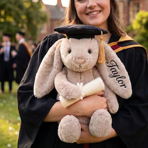 Puede incluir: Una graduada con toga y birrete negros sostiene un conejito de peluche con un gorro de graduación en miniatura y un pergamino. La oreja del conejito está bordada con el nombre "Taylor".