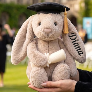Puede incluir: Un conejito de peluche con birrete negro y borla dorada, sosteniendo un diploma enrollado. El conejo es de color marrón claro con el nombre "Diana" bordado en la oreja. El fondo muestra una ceremonia de graduación.