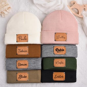 Puede incluir: Una pila de ocho gorros y cintas para la cabeza en varios colores, incluyendo crema, rosa, marrón, gris, verde y negro. Cada gorro y cinta para la cabeza tiene un parche rectangular de cuero con un nombre impreso. Los nombres son Paula, Wallis, Tobias, Robin, Simon, Norton, Blake y Lester.