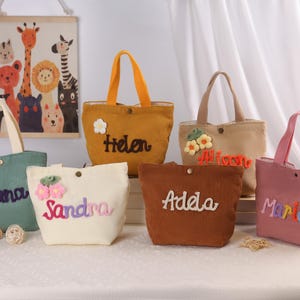 May include: A collection of corduroy tote bags in various colors, each embroidered with a name and adorned with floral embellishments. The bags have matching handles and a button closure. A decorative animal print is in the background.