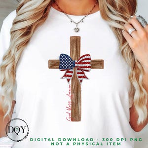 Puede incluir: Camiseta blanca con una cruz de madera estilo acuarela adornada con un lazo patriótico en rojo, blanco y azul, que refleja la bandera estadounidense. Las palabras "God bless America" están escritas verticalmente en la cruz.