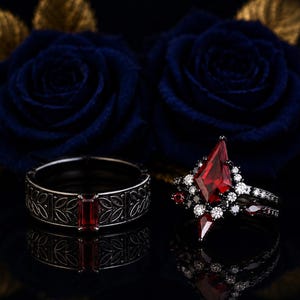 May include: A dark metal band ring featuring a rectangular red gemstone and leaf engravings, paired with a matching engagement ring set with a large kite-shaped red gemstone surrounded by smaller white and black stones on a dark metal band.