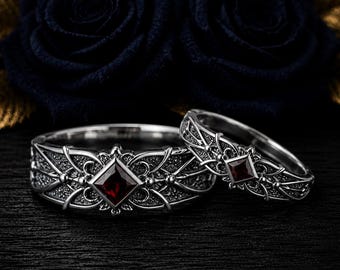 Könnte beinhalten: Zwei silberne Ringe mit filigraner Metallarbeit und einem zentralen, quadratisch geschliffenen dunkelroten Edelstein. Die Bänder zeigen ein detailliertes Gittermuster mit kleinen Metallkugeln und einem tiefroten Stein in der Mitte.