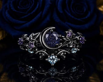 Könnte beinhalten: Ein Ring aus schwarzem Metall mit einem runden, dunkelblauen Sandstein-Cabochon in der Mitte, der von einer Mondsichel eingerahmt wird. Das Band ist mit kleinen violetten und hellblauen Steinen, Sternen und Blattmotiven verziert.