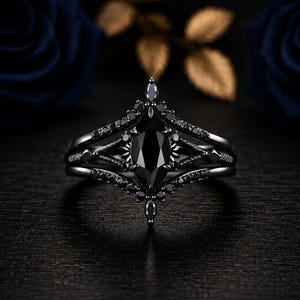 Peut inclure: Une bague en métal noir avec une pierre centrale noire taille marquise entourée de petites pierres noires serties dans un design pointu à plusieurs anneaux. La bague présente une finition sombre polie avec des détails complexes.