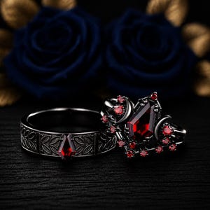 Peut inclure: Un ensemble de bagues en métal noir comprenant un anneau gravé d'un motif de feuilles avec une pierre rouge en forme de cercueil, et une bague assortie ornée d'une grande pierre rouge taillée en cercueil entourée de petites pierres et de lunes.