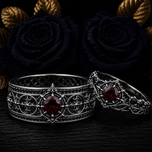 Pode incluir: Dois anéis de metal escuro com padrões intrincados e pequenas pedras pretas. Cada anel é centrado com uma pedra preciosa redonda de cor vermelho-profundo, inserida numa armação pontiaguda em forma de estrela com acabamento escuro.