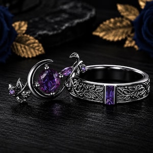 May include: A set of two black metal rings featuring intricate vine and leaf engravings. One ring displays a crescent moon motif with a round purple gemstone, while the other band features a rectangular purple baguette-cut gemstone centred.
