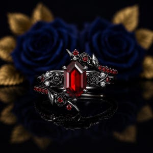 May include: A black metal ring featuring a central hexagonal red gemstone. The band is decorated with sculpted black metal roses and small embedded red stones. The design includes intricate floral details that wrap around the central setting.