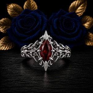 Può includere: Un anello in argento con una gemma rosso scuro centrale a taglio marquise. La fascia in metallo presenta dettagli in filigrana intricati, appuntiti e curvi che si estendono dall'incastonatura. L'anello è posizionato davanti a due rose blu scuro.