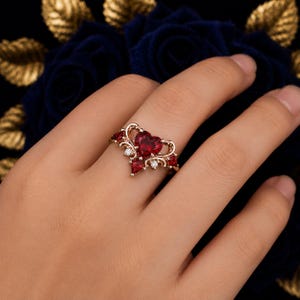 May include: A rose gold ring featuring a central heart-shaped red gemstone flanked by smaller red stones and clear accent stones. The metal band displays intricate scrollwork details that frame the primary heart-shaped red centerpiece.