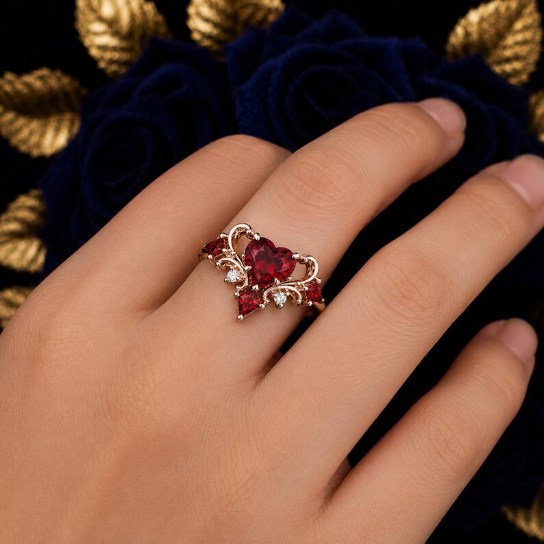 May include: A rose gold ring featuring a central heart-shaped red gemstone flanked by smaller red stones and clear accent stones. The metal band displays intricate scrollwork details that frame the primary heart-shaped red centerpiece.