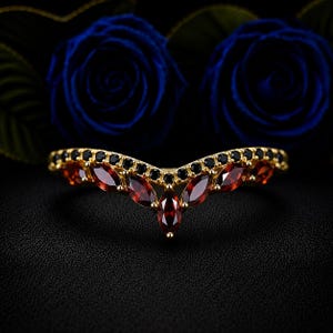 Könnte beinhalten: Ein V-förmiger Goldring mit einer Reihe roter Granat-Edelsteine im Marquise-Schliff am unteren Rand und einer Reihe kleiner runder schwarzer Steine am oberen Rand, die eine Chevron-Silhouette mit einer polierten Metalloberfläche bilden.