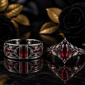 Puede incluir: Un par de anillos de metal oscuro con piedras preciosas de color rojo intenso. Un anillo tiene una banda ancha con una piedra roja central de corte baguette y grabados de hojas. El otro anillo presenta una piedra roja de corte marquesa.