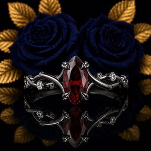 Puede incluir: Un anillo de metal negro con una piedra preciosa roja oscura de corte marquesa sujeta por garras en forma de hoja. La banda tiene un diseño de enredadera, acentuado con pequeñas piedras redondas transparentes y detalles de hojas esculpidas.