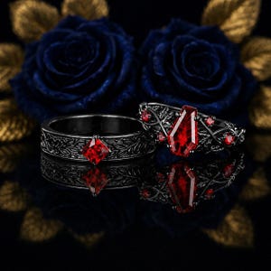Może przedstawiać: Dwa pierścionki z czarnego metalu z misternymi grawerunkami liści. Jeden ma czerwony kamień o szlifie kwadratowym, a drugi czerwony kamień w kształcie trumny otoczony mniejszymi okrągłymi czerwonymi kamieniami w oprawie przypominającej pnącza.