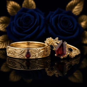 May include: Two gold rings featuring intricate vine and leaf engravings. One ring has a small coffin-shaped red garnet, while the other features a larger coffin-shaped red garnet centerpiece surrounded by small gold roses and black accent stones.