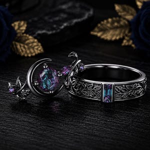 May include: A set of two black metal rings featuring intricate vine and leaf engravings. One ring displays a crescent moon motif with a round, color-shifting purple and blue gemstone, while the other features a rectangular, color-shifting gemstone.