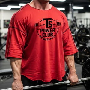 Op de afbeelding: Rood shirt met korte mouwen met de tekst "TS POWER CLUB NO WEAKNESS" in zwart. Het shirt heeft een rond logo met een halterontwerp. De persoon houdt een halter vast in een sportschool.